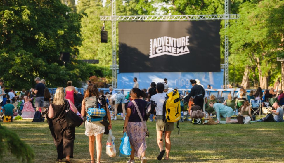People watching an outdoor cinema screen showing "Adventure Cinema" logo on a sunny day in a park.